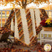 The completed 28th Floor quilt, colored in bold neutral tones like cream, brown, red, and gold, artfully draped over a white porch railing, framed by vibrant fall leaves and garden decor.