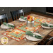 The completed Log Cabin Table Runner colored in autumn-themed prints from the Harvest Blooms collection by Wilmington prints, staged on a rustic wooden table with black chairs, four coordinating place settings with plates and silverware, and a galvanized nickel tin of flowers in the center of the runner.