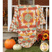 The completed Harvest Blooms Quilt colored in autumn-themed prints from the Harvest Blooms collection by Wilmington prints, artfully draped over a white porch and staged with coordinating fall flowers and pumpkins.