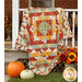 The completed Harvest Blooms Quilt colored in autumn-themed prints from the Harvest Blooms collection by Wilmington prints, artfully draped over a white porch and staged with coordinating fall flowers and pumpkins.