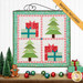 The completed Pieceful Patchwork - December wall hanging depicting Christmas trees and presents in red, blue, green, and white fabrics, hung on a white paneled wall and staged with coordinating décor. A yellow banner in the upper right corner reads "Backing Included!"