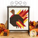 Thanksgiving themed table top banner featuring a Turkey with red, orange, and yellow feathers on a white background, hung on a display hanger and staged with coordinating decor.