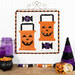 The completed October project featuring two orange spool blocks as trick-or-treat bags with jack-o'-lantern faces and candy, hung on white wall from a craft hanger and staged on a wooden table with coordinating spools and decor.