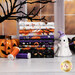 A stack of the fabrics included in the Moonlit Mischief collection, with a window and a dead tree and a Jack-O-Lantern and a ghost and a witches boot and thread.