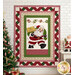 The completed Ribbon Border Panel Quilt colored in red, green, pink, and cream fabrics from the Santa's Coming to Town collection by Henry Glass Fabrics, hung on a white subway tiled wall and staged with a decorated tree and cozy coordinating décor.