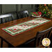 The completed table runner made with log cabin blocks of red, green, mint, and white fabrics from the Mistletoe & Holly collection, laying in the center of a wooden dining table with coordinating décor.