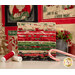 A stack of fabrics included in the Holiday Cheer collection, with a panel, and a tree and a candle and thread and a candy cane.