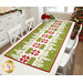 The completed Christmas Village Runner, colored in bright green, white, and red fabrics from the Buon Natalie collection by Moda Fabrics. The project is staged on a white table in a brightly lit kitchen while a decorated Christmas tree sits in the back corner.