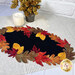The completed Falling Leaves wool mat, a black mat framed by contrasting red, orange, yellow, and brown falls leaves, staged on a table with a lacy tablecloth and a candle.
