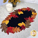 The completed Falling Leaves wool mat, a black mat framed by contrasting red, orange, yellow, and brown falls leaves, staged on a table with a lacy tablecloth and a candle.