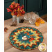 The Point of View Folded Star Table Topper colored in teal, green, cream, gold, and brown fabrics from the Autumn Breeze collection by Northcott fabrics, staged on a wood table with coordinating fall decor and glassware.