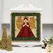 A quilted wall hanging featuring an angel with a golden halo, surrounded by festive patterns and colors, hung on a white display board.
