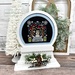 The fully assembled and painted Snow Globe Wood Display, staged on a white tabletop next to a tiny decorative tree, displaying a festive cross-stitch project.
