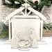 The fully assembled and painted Distressed Nativity Stable Cross Stitch Display, staged on a white tabletop with coordinating festive décor in the background and figurines of Mary, Joseph, and baby Jesus.
