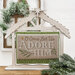 The fully assembled and painted Distressed Nativity Stable Cross Stitch Display, staged on a wooden countertop with coordinating festive décor, displaying a cross-stitch project that says "O Come Let Us Adore Him".