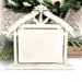 The fully assembled and painted Distressed Nativity Stable Cross Stitch Display, staged on a white tabletop with coordinating festive décor in the background.