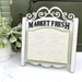 The fully assembled Market Fresh Display with shabby chic white paint and black lettering with a green mount and cream aida cloth on a white table.