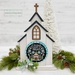 The fully assembled and painted City Chapel Cross Stitch Display Backer, staged on a white tabletop with coordinating garland and winter décor, displaying a wintery cross-stitch project.