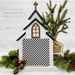 The fully assembled and painted City Chapel Cross Stitch Display Backer, staged on a white tabletop with coordinating garland and a small decorative tree, displaying a small gingham square.