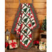 The finished Tree Farm quilt, colored in the rich red and green batiks from the Tonga Mistletoe collection, hung on a rustic wooden ladder and staged in front of a wooden wall with presents and coordinating festive décor framing the quilt.