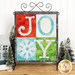 Christmas themed table top banner featuring a grid of four squares with an appliqué snowflake and letters to spell "Joy" in festive colors, hung on a display hanger and staged with coordinating decor.