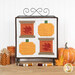 The completed Sew Thankful wall hanging, two spools with pieced leaves paired with two pieced pumpkins in bold autumn colors, hung on a display stand and staged with coordinating spools of thread and autumn decor.
