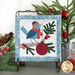 The completed On Wander Lane Mini Quilt for December, an appliqué bird hanging a Christmas ornament on a tree, with hand stitched details. The project is staged on a neutral table beside coordinating Christmas decor.