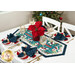 The completed Easy Striped Table Runner Kit colored in teal, white, blue, and red fabrics from the Woodland Wishes by Northcott Fabrics, staged on a white table with coordinating wine glasses, table settings featuring the coordinating napkins, and a basket of red poinsettias in the center.