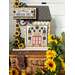 A decorative cross stitched house sign labeled Sunflower Street surrounded by yellow sunflowers and a woven basket.