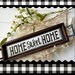 A wooden sign frames a cross stitched Home Sweet Home project, surrounded by decorative flowers on a checkered background.