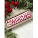 A decorative cross stitch sign saying SANTA CLAUS, framed in wood with festive red accents and greenery.