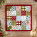 The completed Fusible Grid Hot Pad colored in red, green, white, and light blue fabrics from the Emmitt and Ivy collection by Moda Fabrics. The project is staged on a wood table where a wooden spoon and green garland peek into frame.