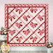 The completed Foundation Paper Piecing Heart & Lattice Quilt colored in pink, red, and white fabrics from the Sew In Love collection by Riley Blake Designs, hung on a pink shiplap wall and staged with coordinating white buckets of pink and white roses.