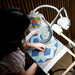 A person stitches a colorful quilt pattern under a magnifying lamp in a well-lit workspace.