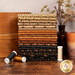 A stack of the fabrics included in the Midnight Cheddar collection with boxes and a candle and a plant and a bottle and thread.