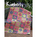 A colorful quilt draped over a wooden bench surrounded by greenery, titled Kimberly.