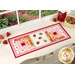 The completed Sew Cute Runner - June table topper, staged on a white dresser, accented with scattered red strawberries, and a red vase sitting in the upper right corner.