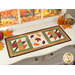The completed Sew Cute Runner - November table topper, staged on a white dresser, accented with scattered leaves, a pumpkins and a drink of apple cider in the upper right corner. 