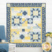 The completed Prayers of the Heart Quilt, colored in dark blue, blue and yellow fabrics from the Middleboro Road collection. The quilt is displayed on a white paneled wall and staged with coordinating flowers and a blue knit blanket draped over a chair.