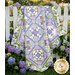 The completed Bouquet of Hydrangeas Quilt, colored in powder blue, purple, green, and white fabrics from the Hydrangeas in the Garden collection from Riley Blake Designs, artfully draped over a white picket fence at sunset among full bloom hydrangea flowers.