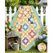 The completed Churn Dash Layer Cake Quilt colored in vibrant prints from the Aunt Grace Goes Scrappy collection by Marcus Fabrics, draped over a white picket fence and surrounded by coordinating bunches of flowers.