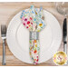 A top down shot of one of the napkins with the rainbow floral fabric faced out, staged on a white plate with cutlery on a wooden table.