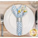 A top down shot of one of the napkins with the light blue gingham fabric faced out, staged on a white plate with cutlery on a wooden table.