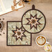 The finished Square and Round Folded Star Hot Pads made with the Coffee Time collection in sage, featuring chocolate brown and cream stars at the center of the hot pads surrounded by sage green fabric with coffee related phrases. The hot pads are staged on a wooden table with coordinating décor, a gold shabby fabrics logo is in the bottom right corner.