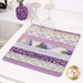 A colorful patchwork drying mat in various shades of purple, placed beside two glasses and a sink.
