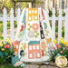 The completed Spring Town Quilt colored in vibrant fabrics from the Delightfully Yours collection by Moda Fabrics, artfully draped over a white picket fence among flowers and garden items.