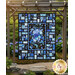 The completed Stained Glass Panel Quilt colored in the bright blue fabrics of the Escapade collection, with a stunning floral panel at the center, contrasted with striking black borders. The quilt is hanging from a wooden pergola set in a peaceful garden of blue hydrangeas with a gold Shabby Fabrics logo in the bottom right corner of the image.