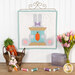 The completed April spool, featuring a large gray bunny holding a blue spool with an orange carrot, hanging on a white wall above a wooden table with coordinating spools and décor.