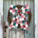 A rustic door features a woven wreath adorned with a colorful patchwork bow in red, green, and white.