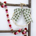 A green and white plaid bow hangs from a wooden hanger, adorned with wooden beads and a garland of felt balls.